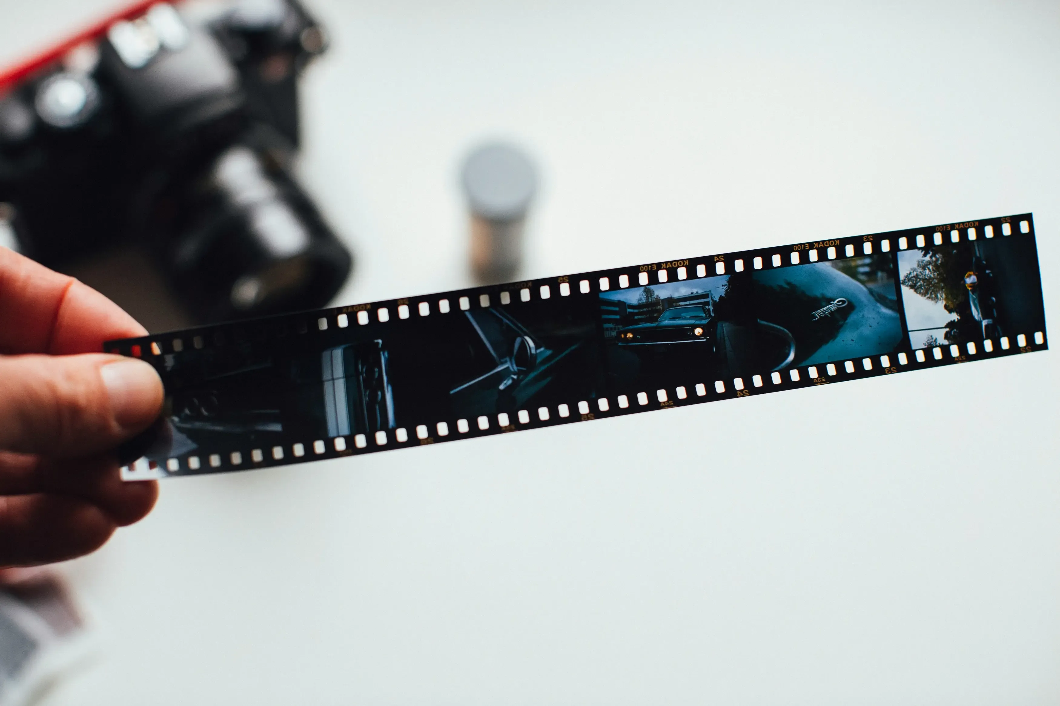 A strip of developed 35mm film held up to the light, a camera on the table behind it.